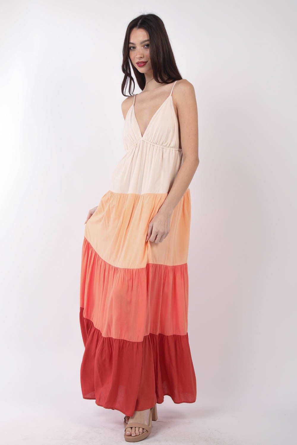 A woman stands, wearing the VERY J Color Block Tiered Maxi Cami Dress in shades of beige, peach, and red, paired with beige sandals. Her right hand holds the side of her dress as she poses against a plain white background—a truly fashionable choice.