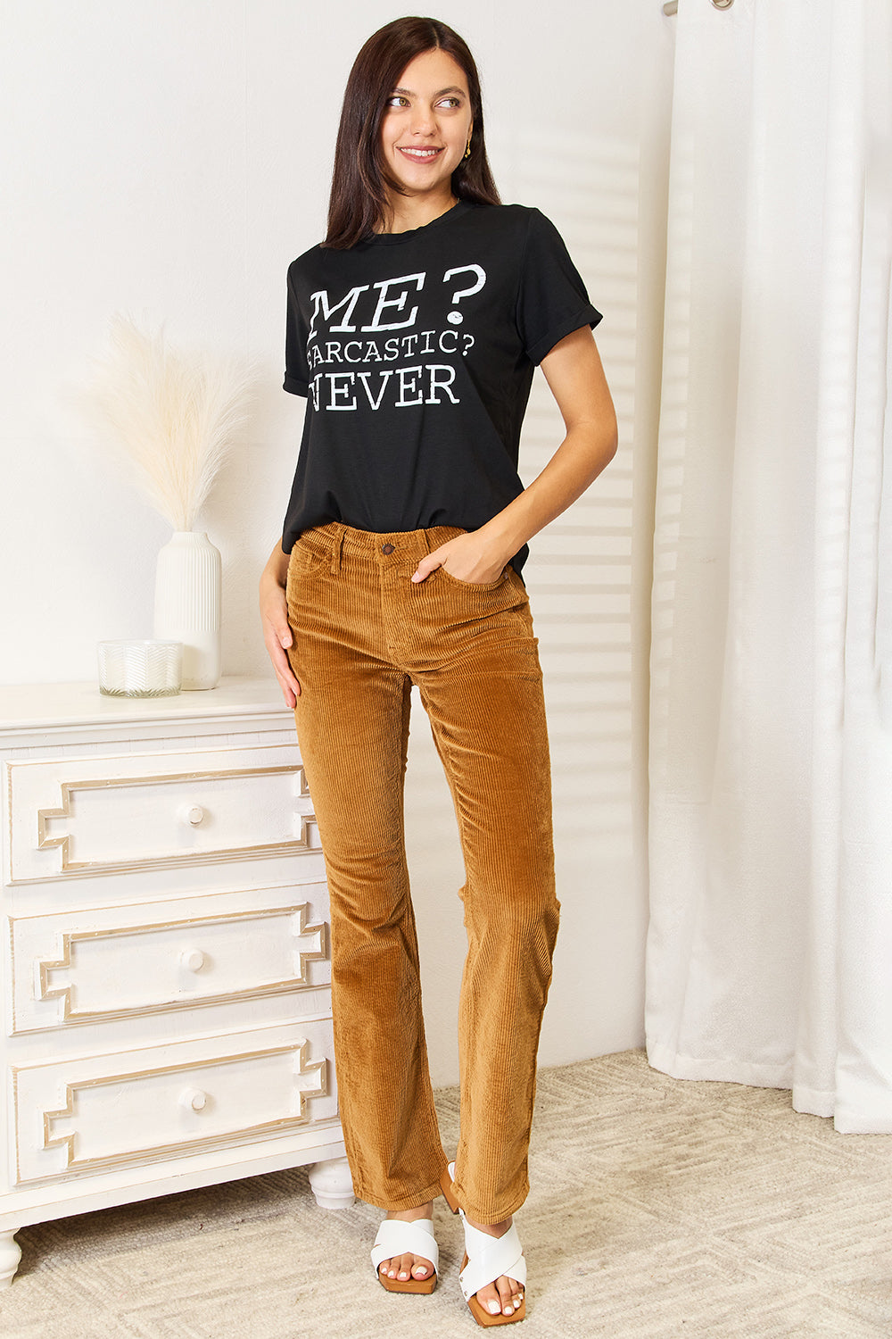 A woman with long dark hair smiles while wearing a trendy black Simply Love Letter Graphic Round Neck T-Shirt featuring the text "ME? Sarcastic? NEVER" and brown corduroy pants. She stands in a room with white walls and decor.