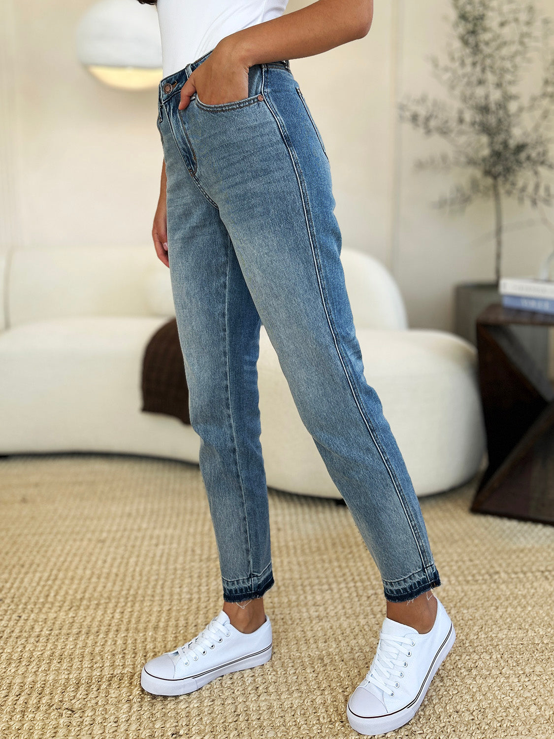 Person wearing a black top, Judy Blue Full Size Mid Rise Rigid Magic Release Hem Jeans, and black sneakers with white laces, standing on a beige carpet with hands in pockets.