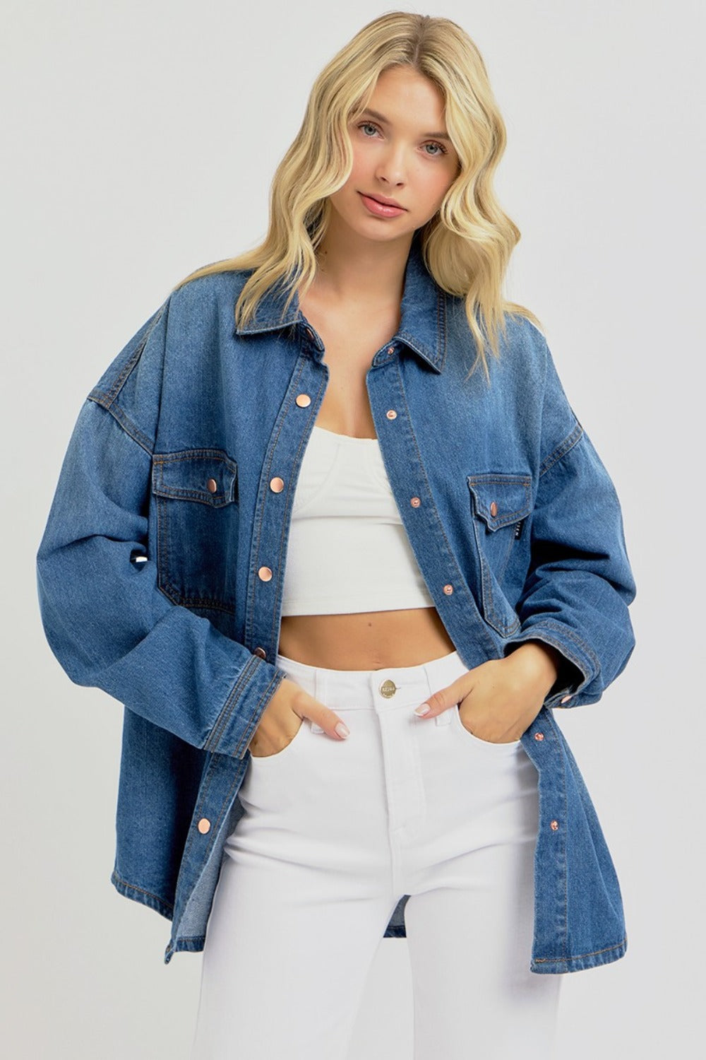 A person with long blonde hair, wearing the RISEN Full Size Snap Down Long Sleeve Denim Shacket over a white crop top and white pants, stands against a plain background.