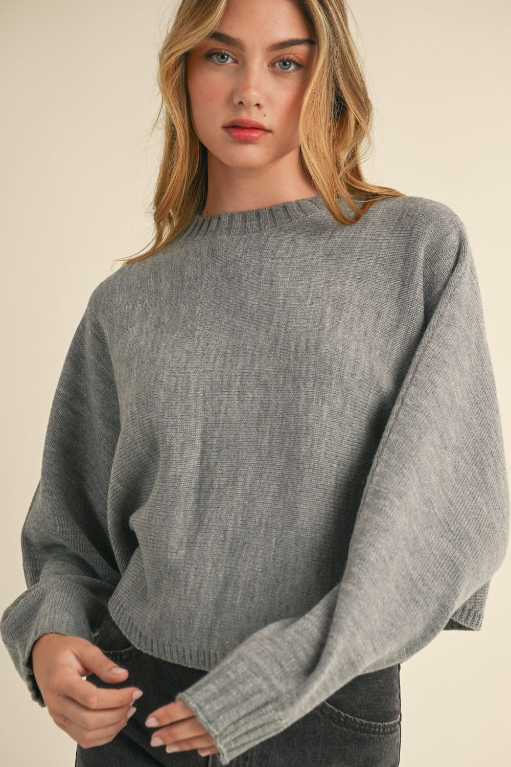 A person with long blonde hair wears the Mable Round Neck Dolman Sleeve Cropped Sweater in grey while standing against a neutral background.