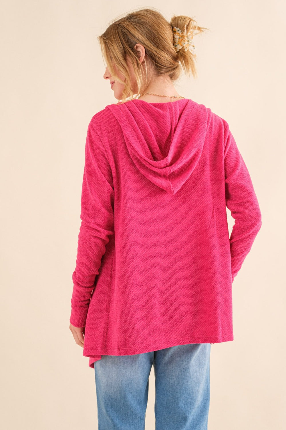 A person wearing the And The Why Full Size Thermal Hooded Open Front Cardigan with Pockets in bright pink over a beige top and blue jeans stands against a plain background. They are smiling and holding the edge of the warm thermal fabric cardigan with one hand.