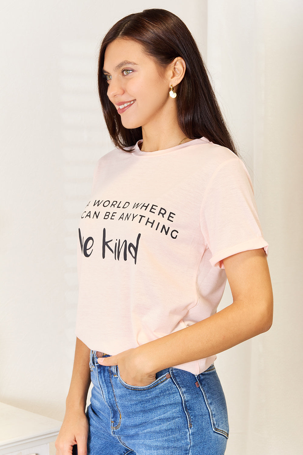 A person wears a Simply Love Slogan Graphic Cuffed T-Shirt in light pink, adorned with the inspiring message, "In a world where you can be anything, be kind," and featuring stylish cuffed sleeves.