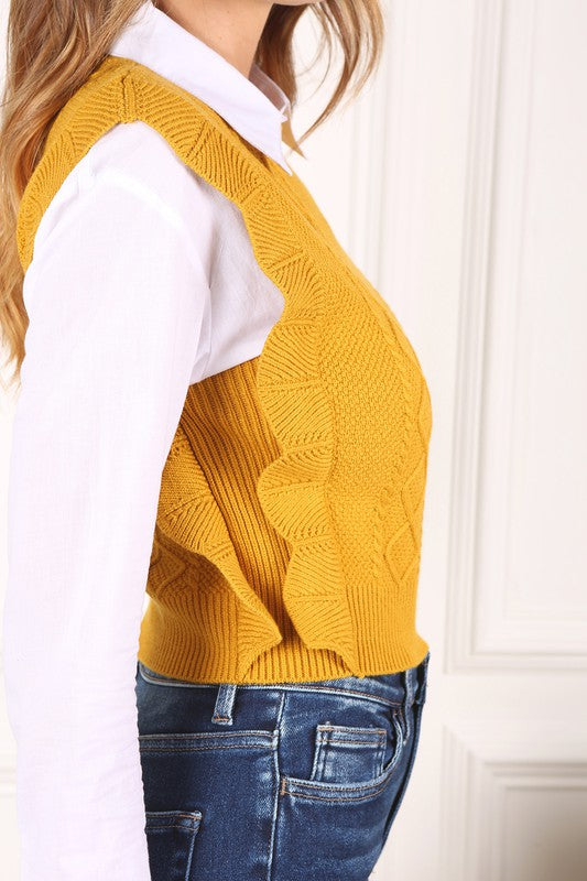 A person wearing a Ruffle sweater vest over a white collared shirt and blue jeans stands against a white-paneled background.