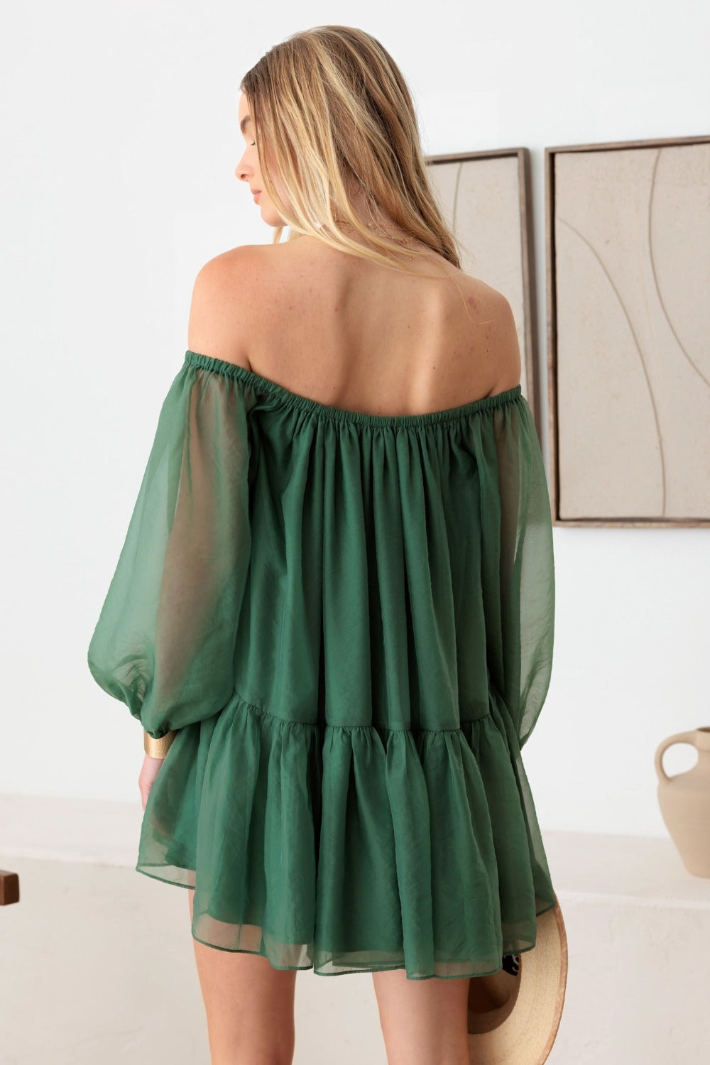 A woman stands indoors near a chair and a framed artwork on the wall, wearing the stunning Le Lis Organza Off Shoulder Puff Sleeve Mini Dress in green. This off-the-shoulder dress with long sleeves makes for a perfect special occasion ensemble.