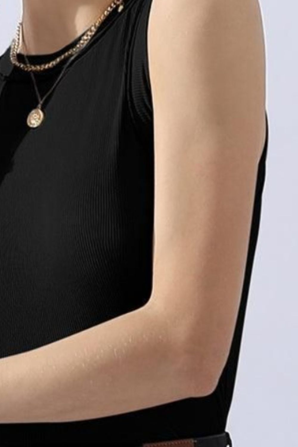 A person wears a versatile and comfortable Ninexis Ribbed Round Neck Tank in black, paired with black jeans and gold jewelry, smiling against a white background.