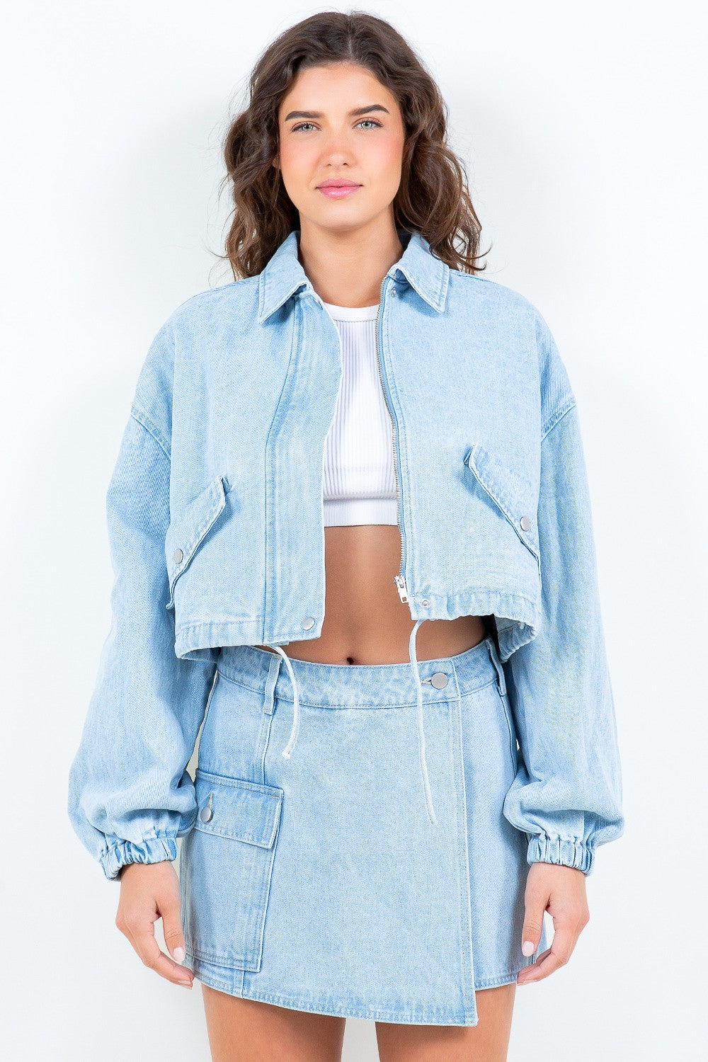 A woman wearing an American Bazi Drawstring Waist Crop Denim Jacket over a white top and a matching light denim skirt stands facing forward against a plain background.