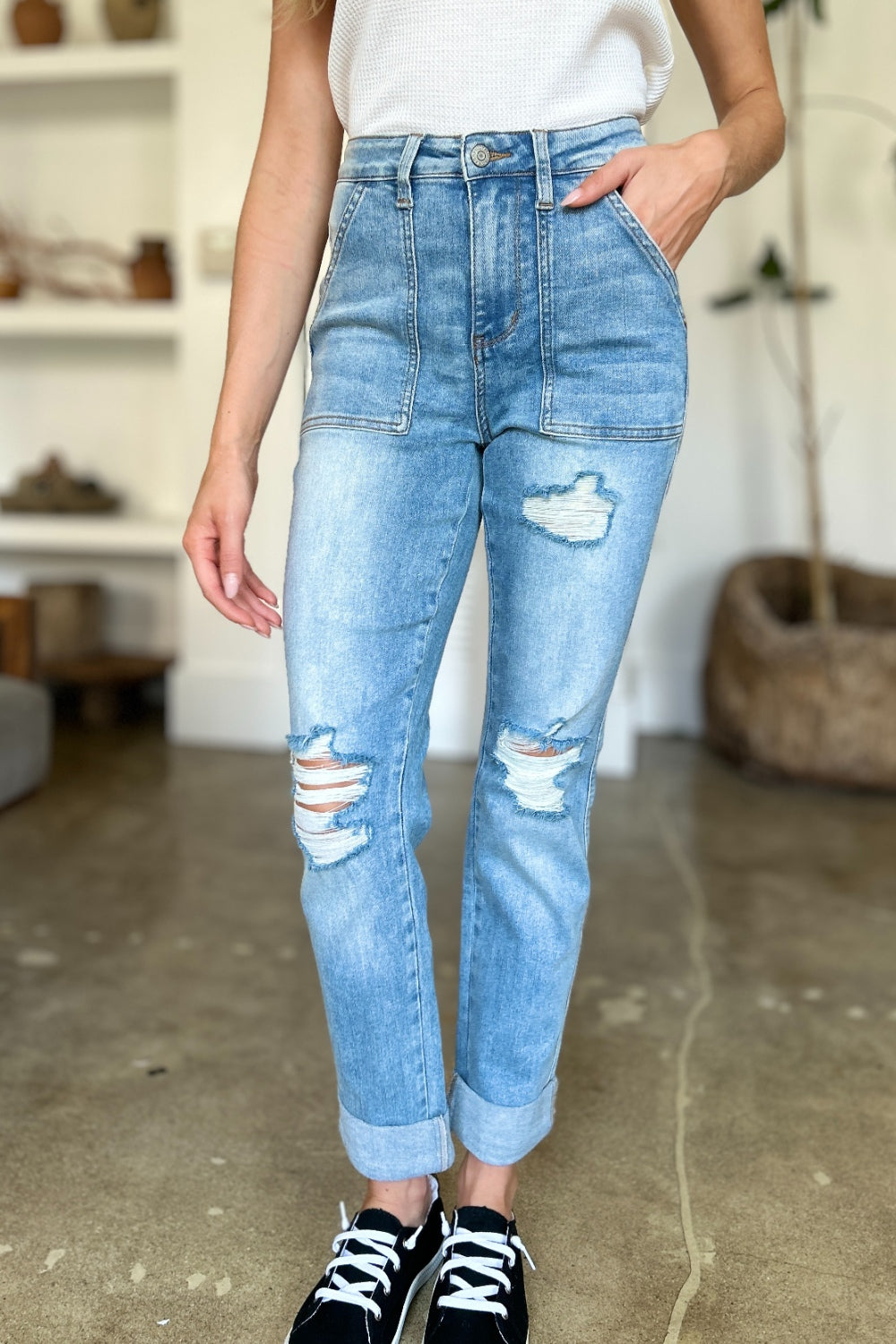 Two women are standing side by side, both clad in black and white tank tops and Judy Blue Full Size Distressed Straight Jeans with Patch Pockets, smiling at the camera in a modern living room.