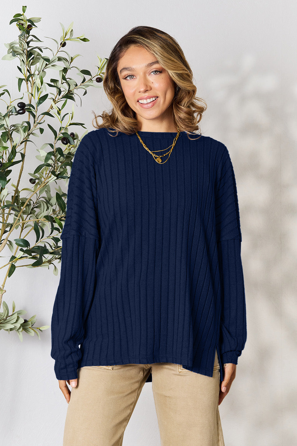 A person wearing the Basic Bae Full Size Ribbed Round Neck Slit Knit Top in blue and beige pants stands and smiles in front of a leafy plant.