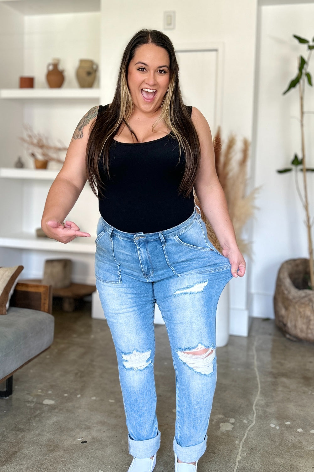 Two women are standing side by side, both clad in black and white tank tops and Judy Blue Full Size Distressed Straight Jeans with Patch Pockets, smiling at the camera in a modern living room.