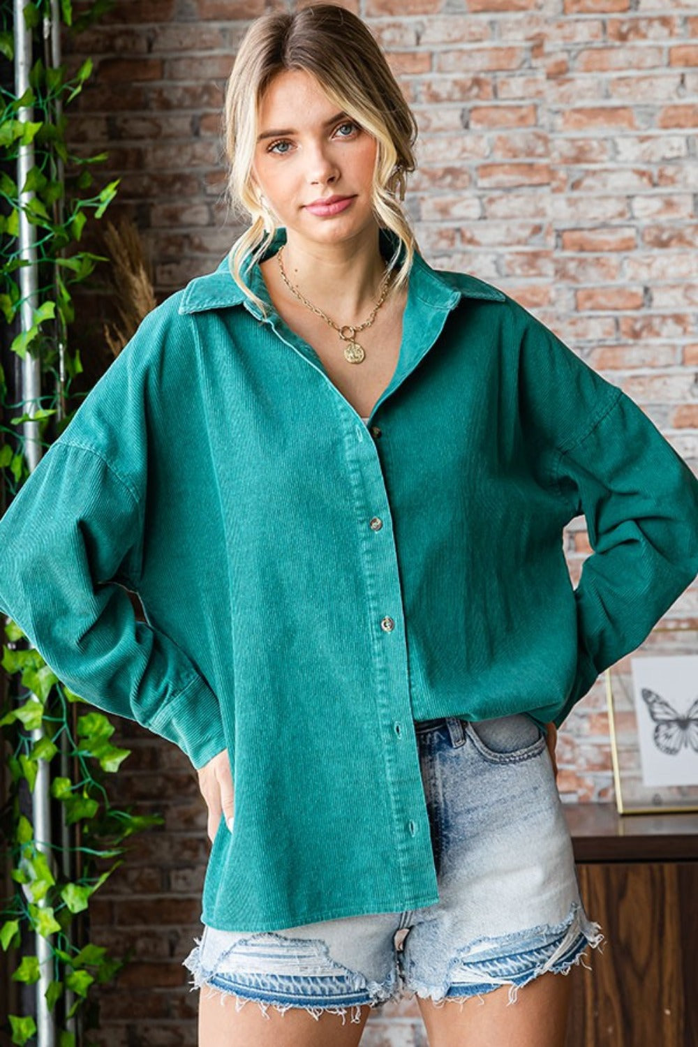 A woman wearing the First Love Washed Corduroy Button Down Long Sleeve Shirt in green, a white top, and denim shorts poses indoors. She stands in front of a brick wall with a leafy plant on the side.