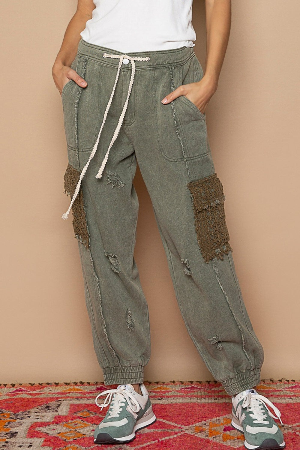 A person sporting POL Distressed Cargo Denim Joggers with Crochet Pockets in green and a white top stands casually on a colorful rug, hands tucked in pockets.