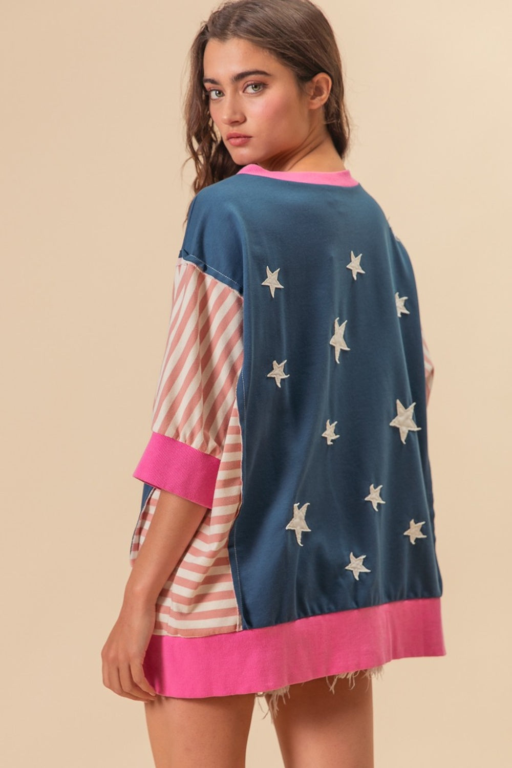 A person wearing a BiBi US Flag Theme Color Block Star Patch T-Shirt, characterized by its oversized fit, blue base with white star patterns, and pink-striped sleeves, stands against a plain background. They have their hand touching their hair.