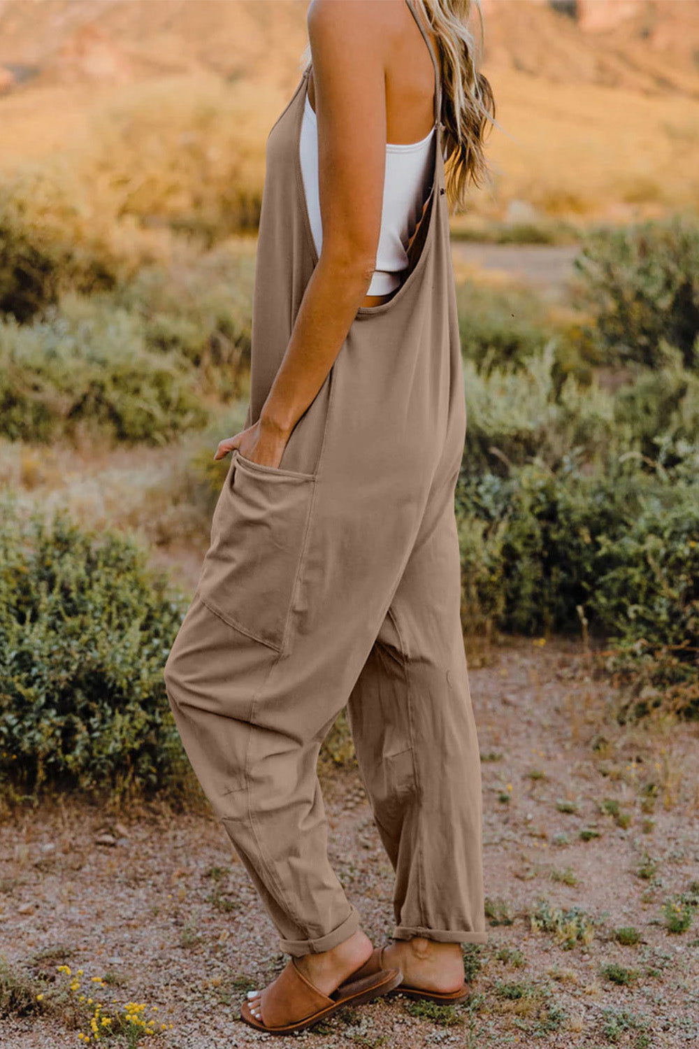 Person wearing a Double Take Full Size V-Neck Sleeveless Jumpsuit with Pockets and sandals, walking outdoors on a dirt trail with bushes in the background.