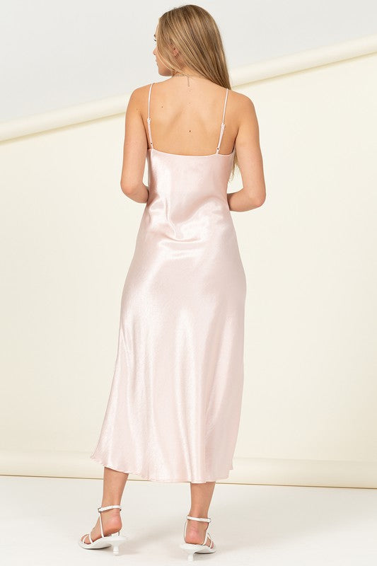 Dressed in the Love Tree Sleeveless Maxi Dress, a woman showcases effortless elegance as she holds the light pink satin skirt to reveal a thigh-high slit, set against a plain background.