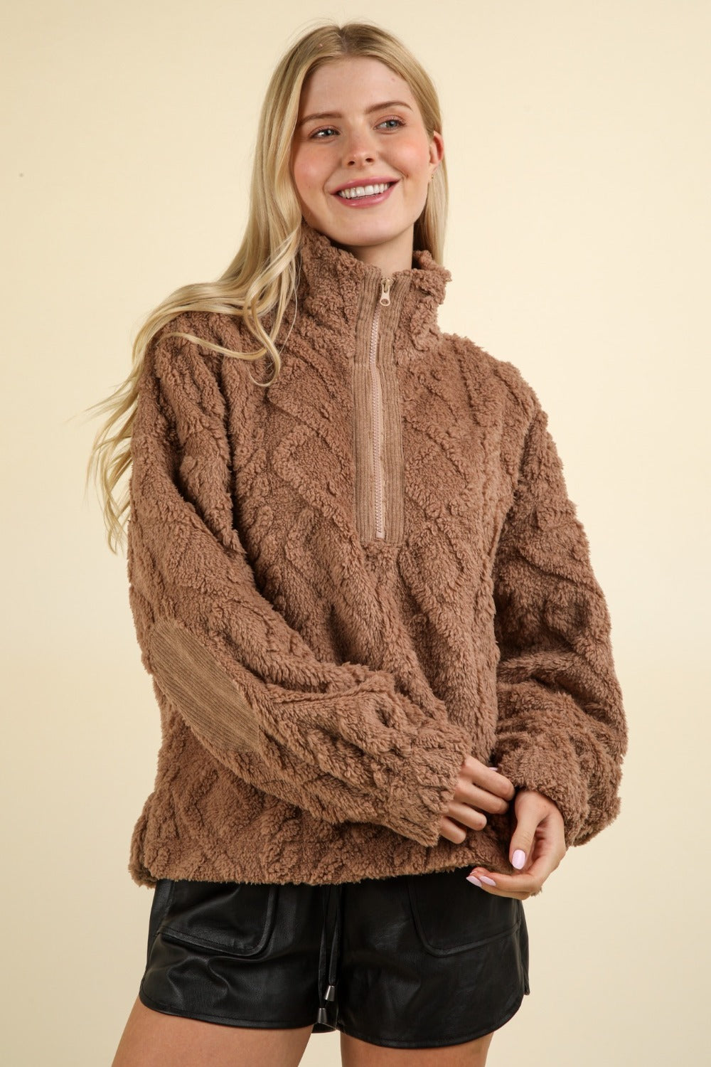 A young woman with long blonde hair is wearing a VERY J Fuzzy Fleece Half Zip Cable Pattern Sweatshirt in brown and black shorts, smiling and facing slightly to the side against a plain beige background.