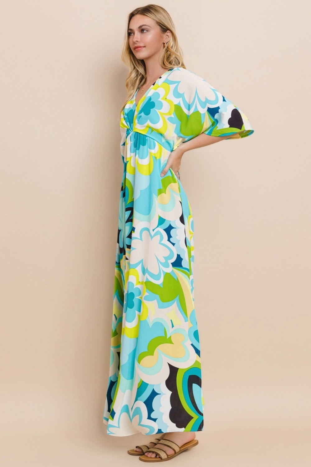 Against a simple backdrop, a woman models the ODDI Floral Printed Slit Maxi Dress, exuding a sense of versatile elegance. This maxi dress embodies summer chic with its vibrant floral design featuring shades of blue, green, and yellow.