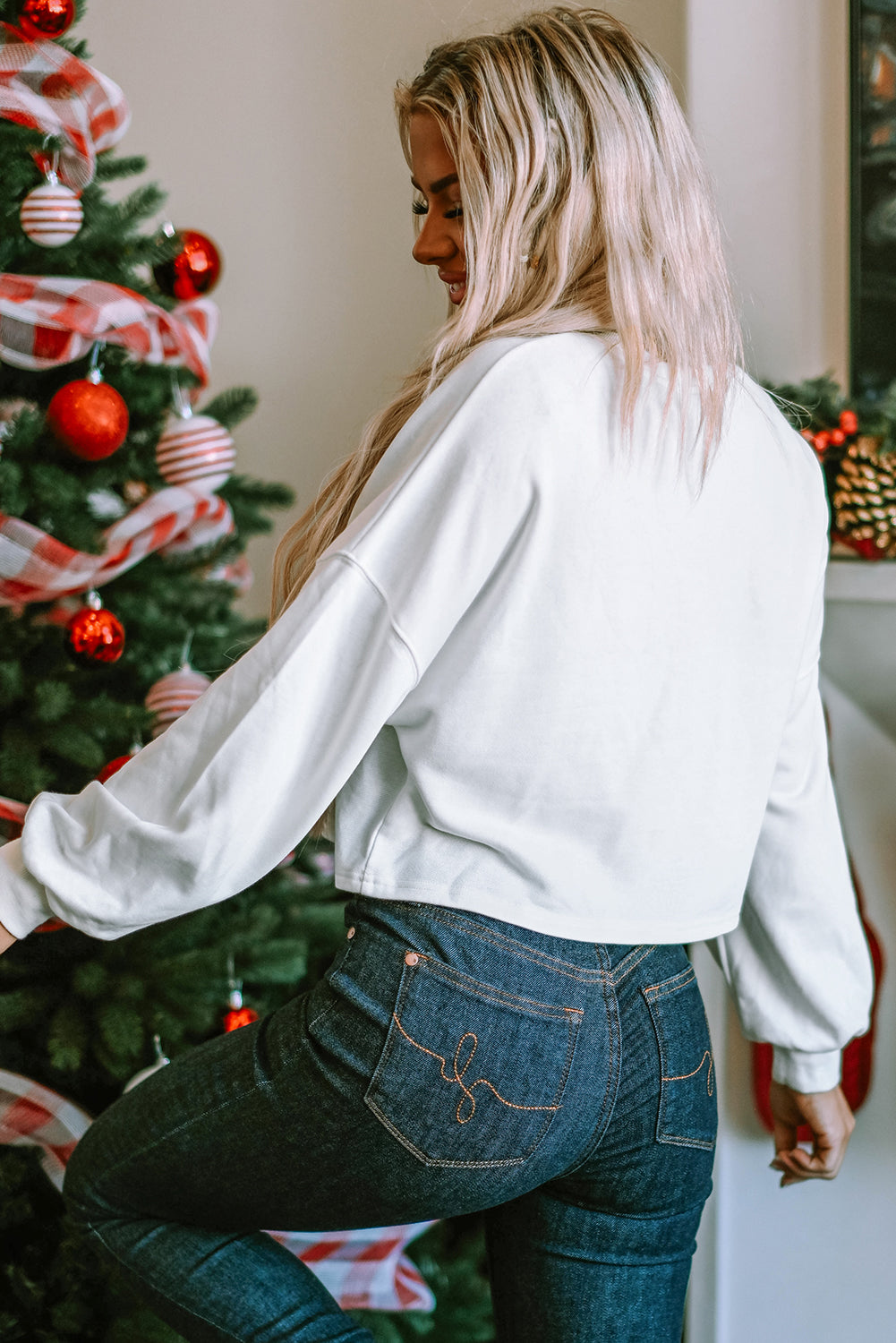 A woman with long blonde hair dressed in a chic White Game Day Lettering Rugby Football Notched Neck Sweatshirt and blue jeans stands near a Christmas tree adorned with red and white ornaments and ribbon.