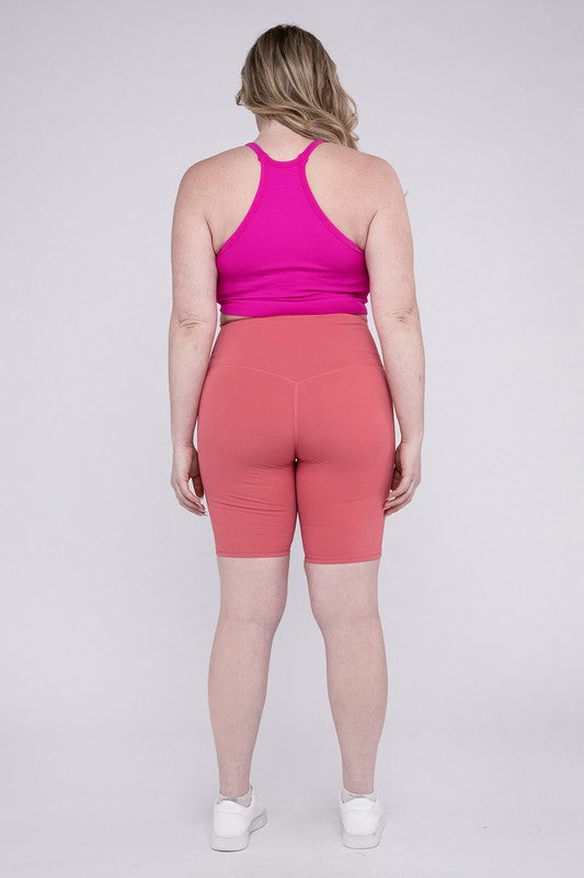 Against a plain background, a person showcases the Plus Athletic High Rise Biker Shorts in coral, crafted from high-quality athletic fabric and paired with a pink top.