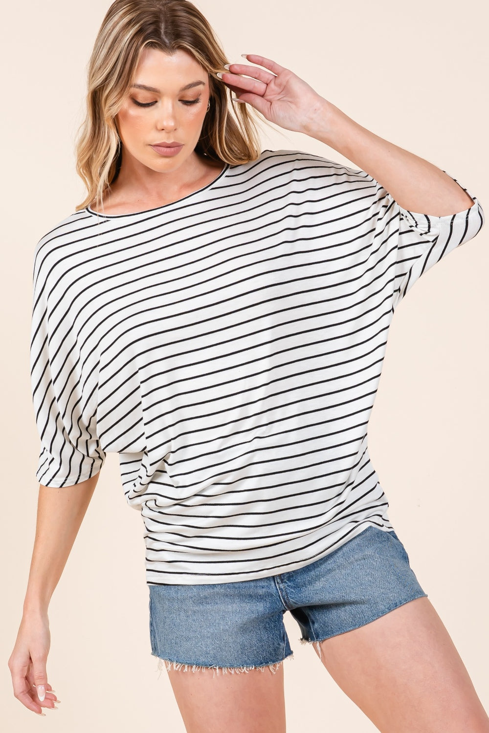 A person is wearing the BOMBOM Striped Boat Neck Dolman Sleeve Top paired with denim shorts, effortlessly embodying a wardrobe staple. The person has their right hand touching their hair and is looking down, against a plain, light-colored background.