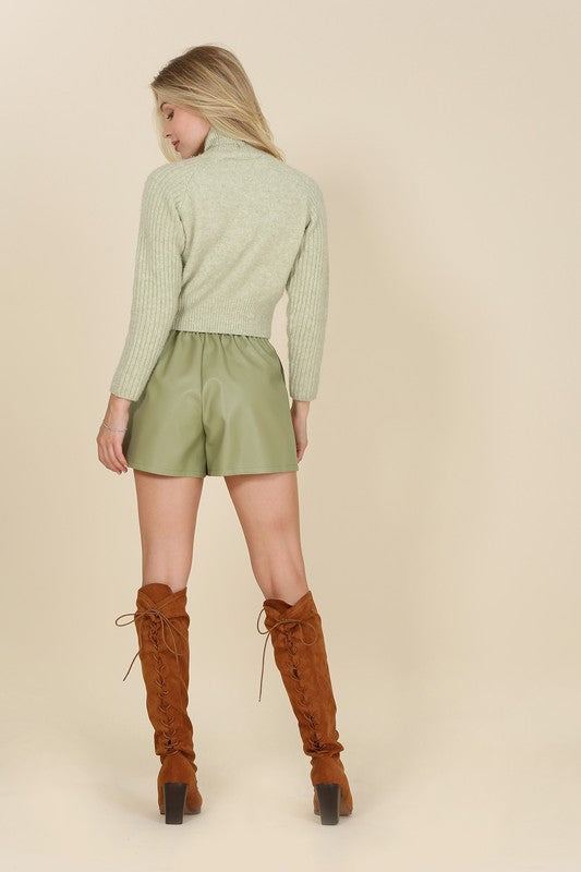 An individual is dressed in a light green sweater paired with pleated vegan leather shorts, featuring a functional pocket where one hand rests.