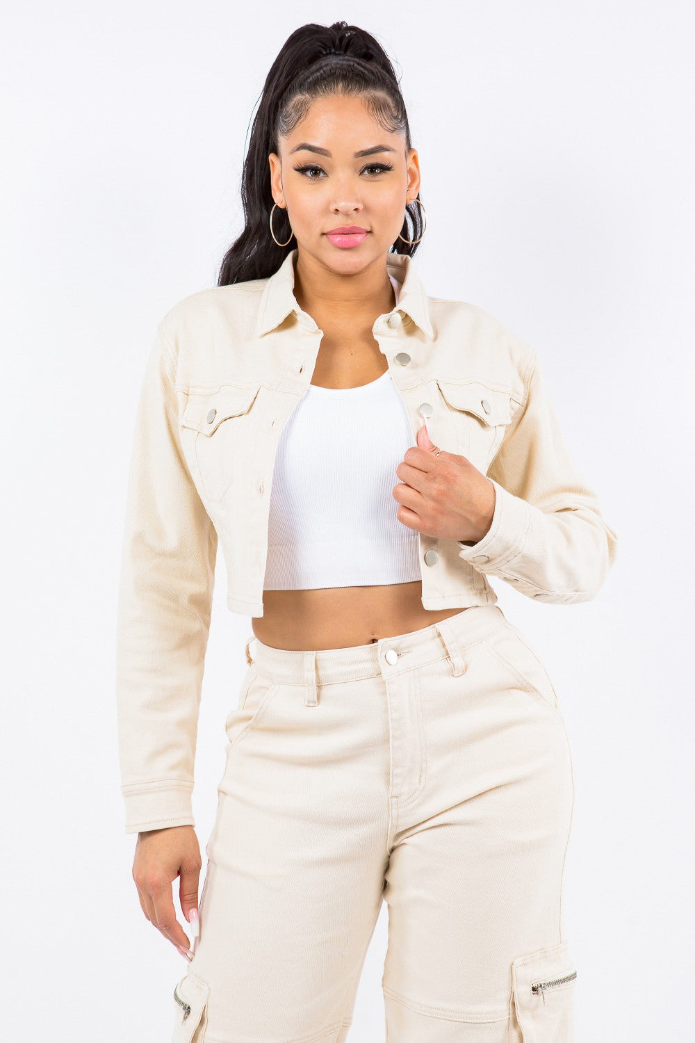 A person wearing an American Bazi Laced Back Cropped Jacket in beige with matching pants and a white crop top stands against a plain white background. They have long dark hair tied in a high ponytail and are looking towards the camera.