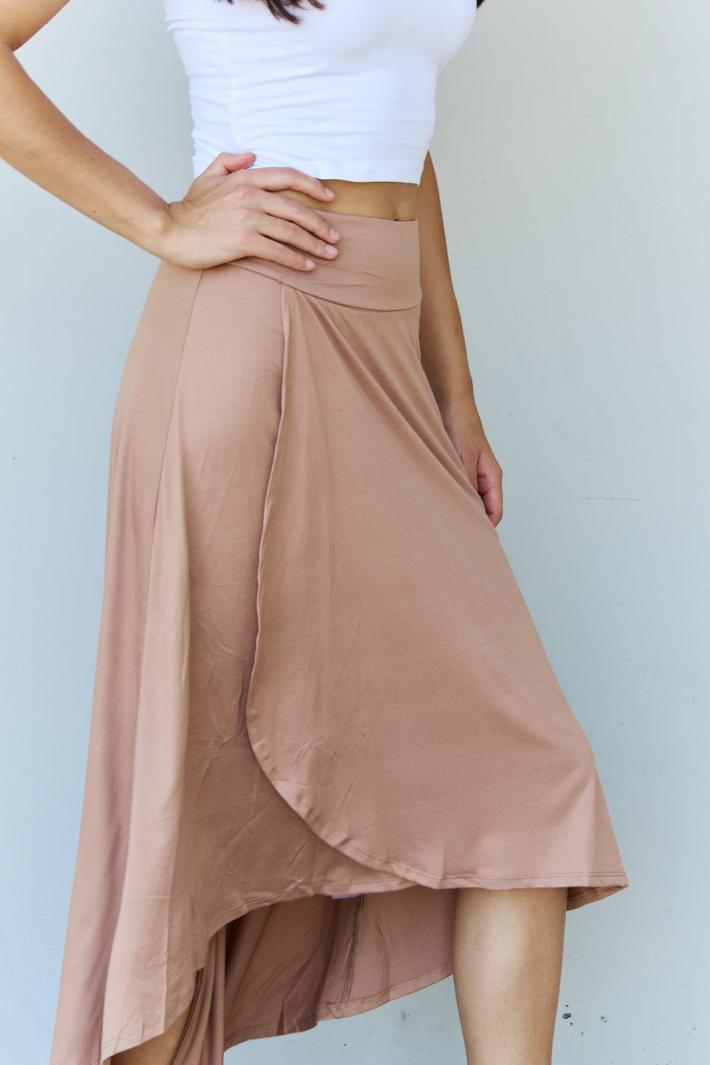 Person wearing a white crop top, the Ninexis First Choice High Waisted Flare Maxi Skirt in Camel with an asymmetrical hemline, and black sandals, standing against a light-colored wall.