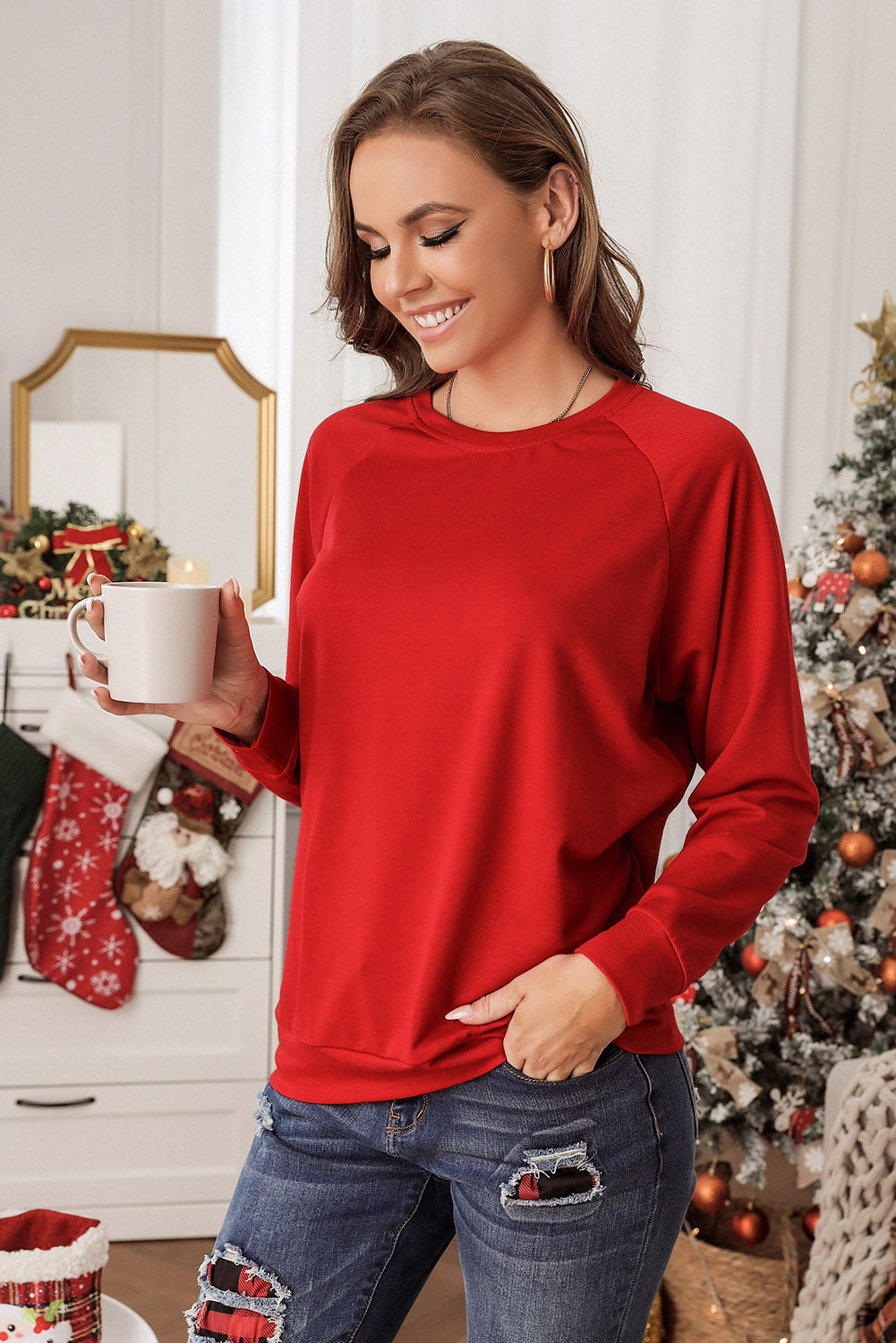 A person wearing a cozy Fiery Red Solid Round Neck Raglan Sleeve Sweatshirt and jeans stands indoors with a decorated Christmas tree and wrapped presents in the background.