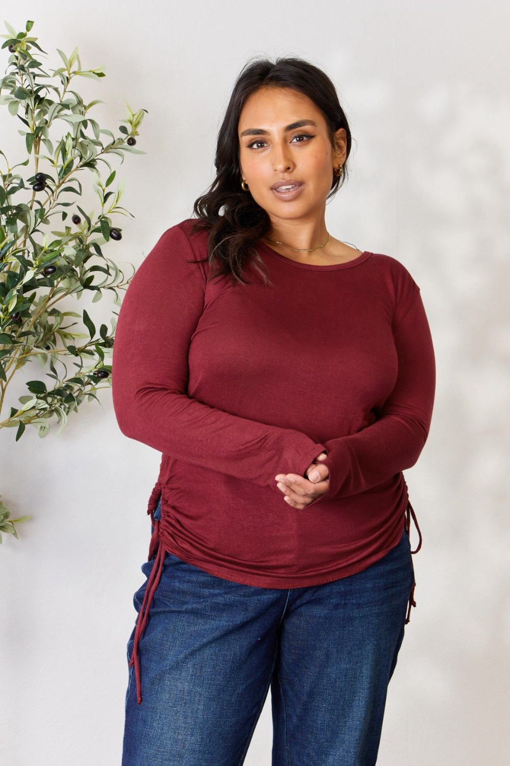 A woman stands in front of a plain background and a small tree, wearing the Culture Code Full Size Drawstring Round Neck Long Sleeve Top in maroon and blue jeans.