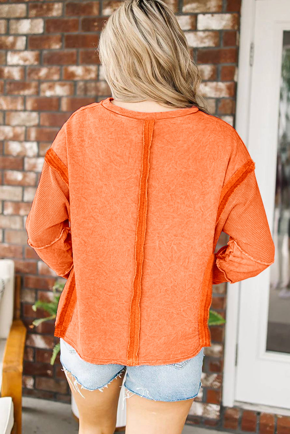 A person with long blonde hair is standing with their back to the camera, wearing an Orange Exposed Seamed High Low Raw Edge Sweatshirt and denim shorts, in front of a brick wall.