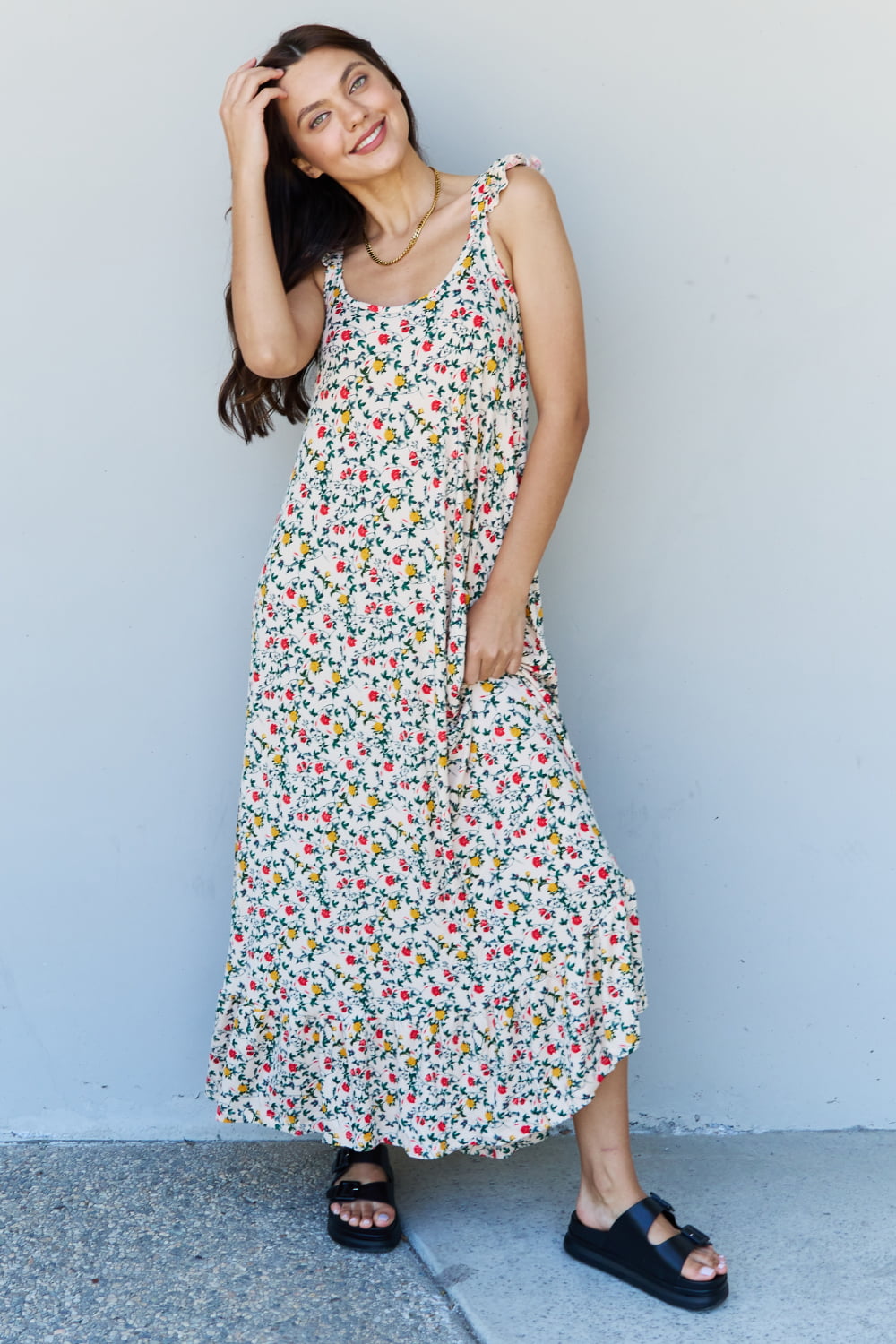 A woman is standing against a gray wall, wearing the Doublju In The Garden Ruffle Floral Maxi Dress in Natural Rose and black sandals, with her left hand raised to her head.