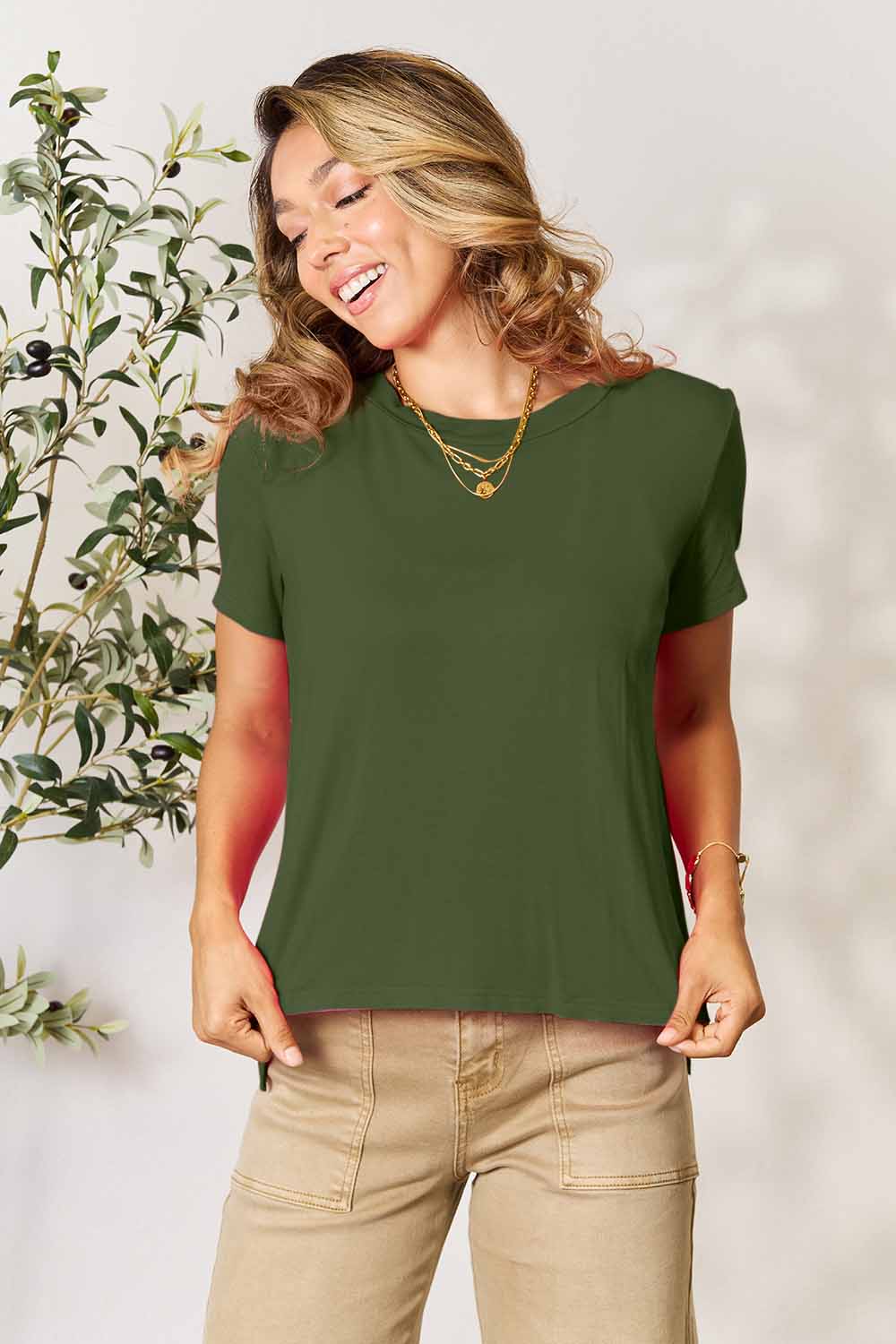 A woman stands next to an indoor plant, smiling, with one hand in her khaki pants pocket. In her pink Basic Bae Full Size Round Neck Short Sleeve T-Shirt and gold jewelry, she radiates casual elegance.