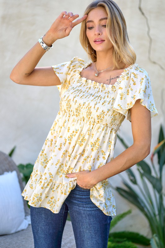 A woman stands outdoors donning a Floral Printed V-Neck Ruffle Top paired with jeans, set against a neutral background, with plants in the foreground, highlighting her fashion-forward wardrobe.