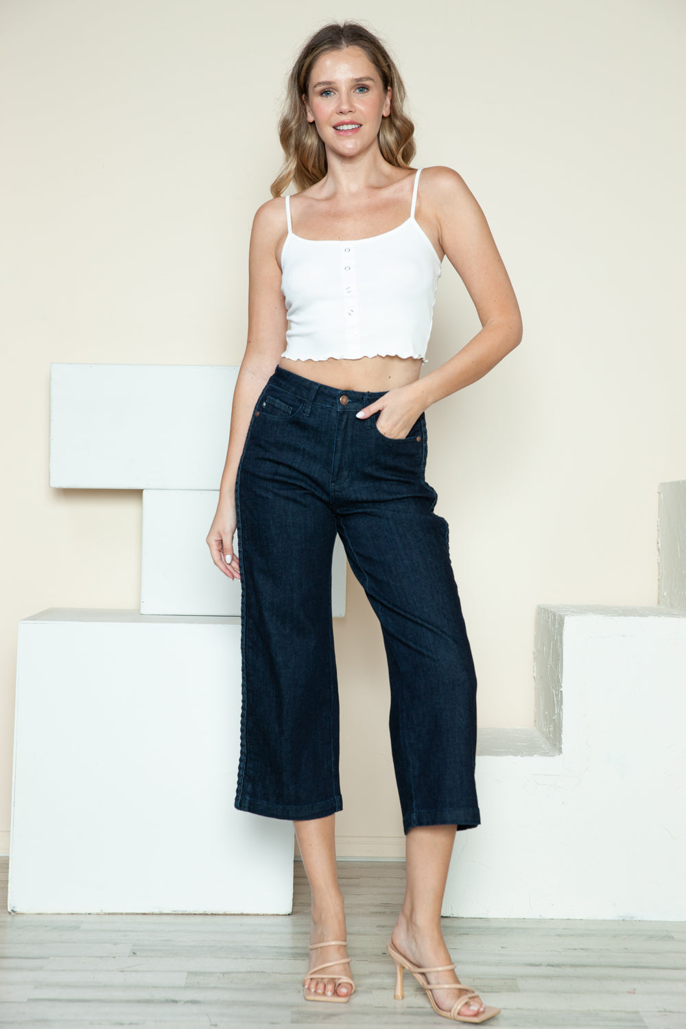 Person wearing Judy Blue Full Size Side Seam Braid Detail Crop Wide Leg Jeans and beige strappy heels, standing on a wooden floor with a white background.