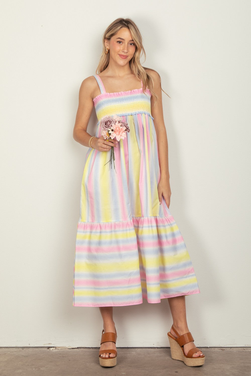 A woman in the VERY J Striped Woven Smocked Midi Cami Dress, featuring pastel stripes, holds a small bouquet of flowers. She pairs the dress with tan platform sandals that complete her chic summer look and stands against a plain white background.