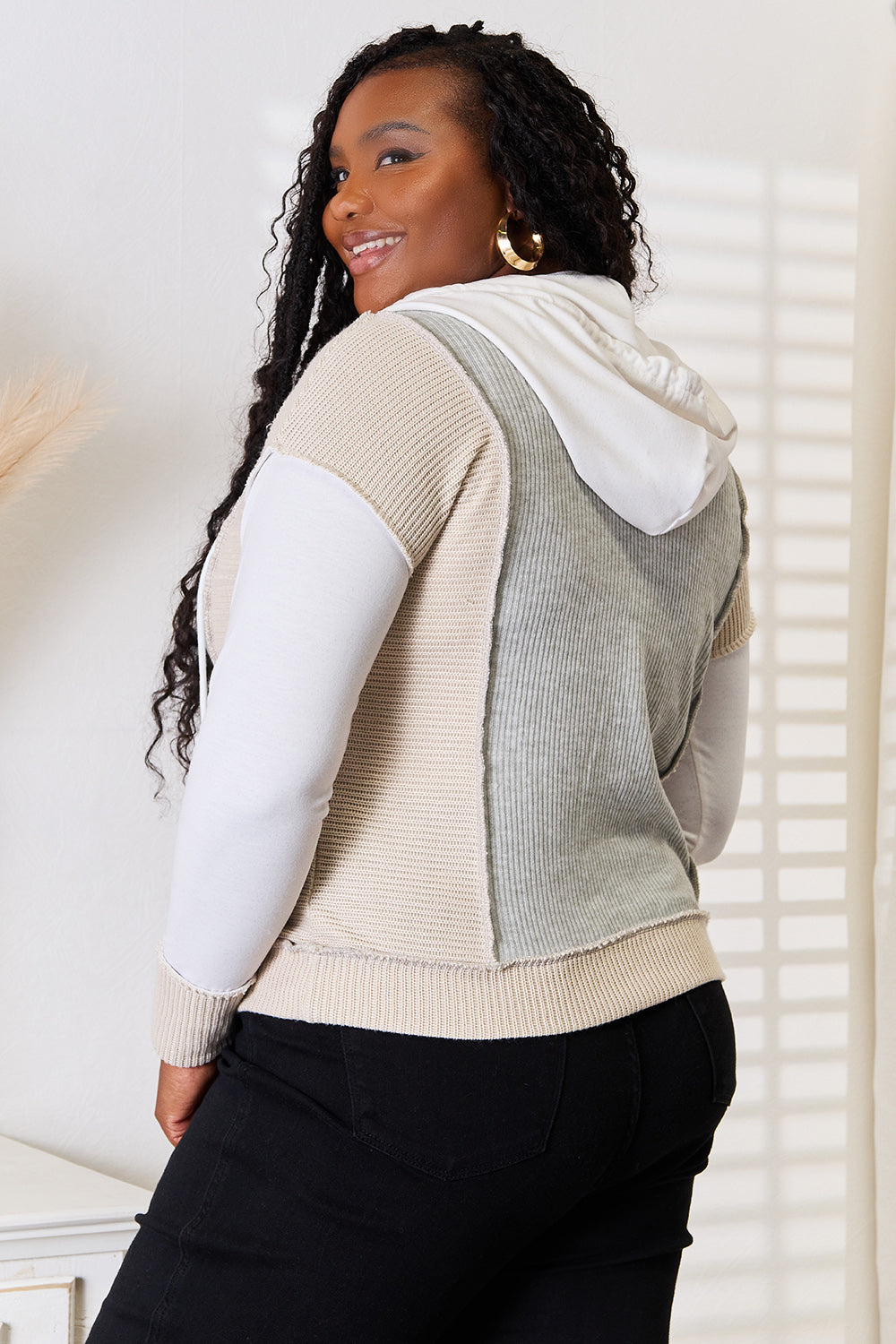 A woman wearing the Double Take Color Block Exposed Seam Drawstring Hoodie poses indoors. She has long braided hair, is smiling, and rests her hand on her hip.