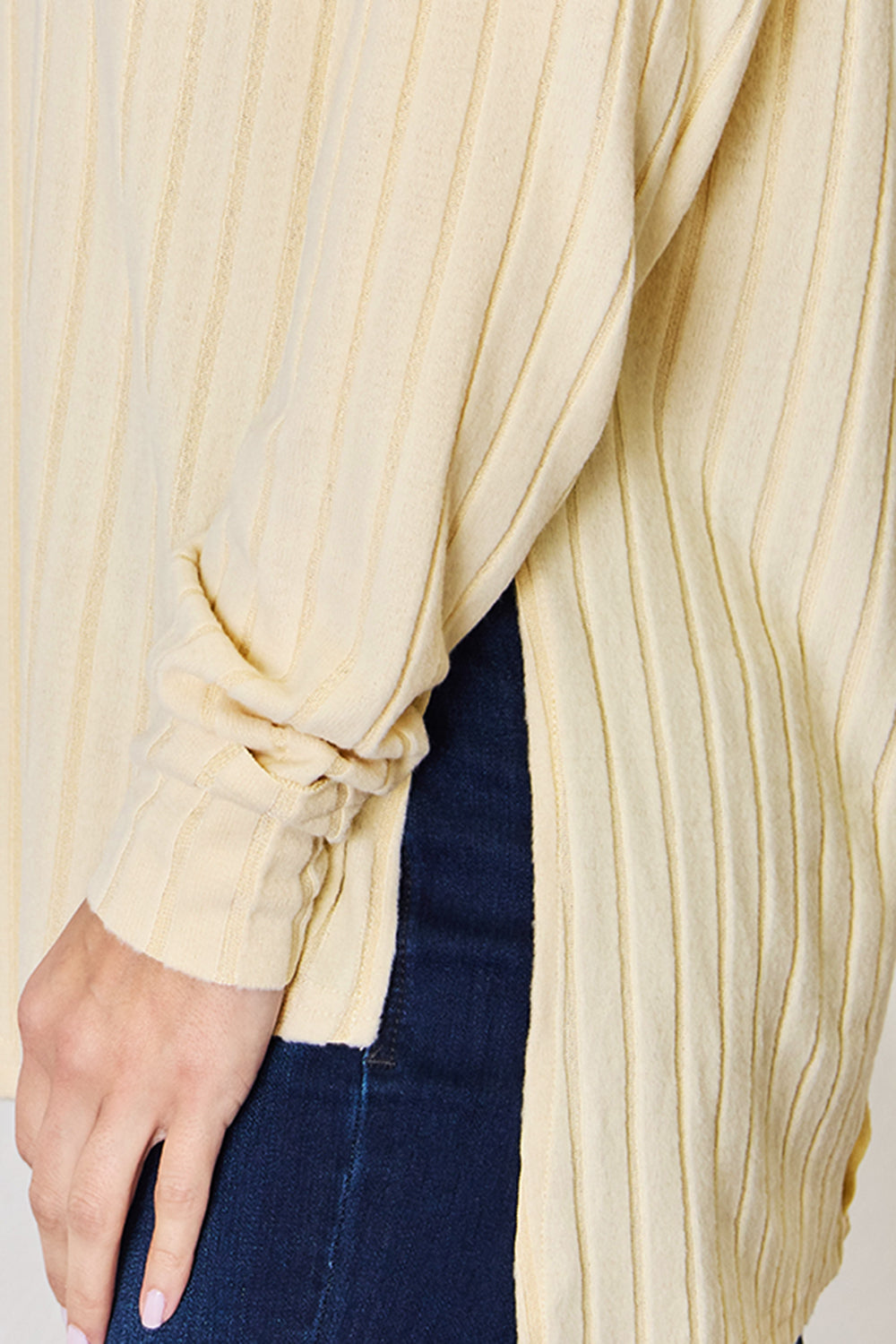 A woman with long blonde hair is wearing a light grey, slightly stretchy, long-sleeve Basic Bae Full Size Ribbed Round Neck Slit T-Shirt and dark blue jeans. She is standing against a light-colored background.