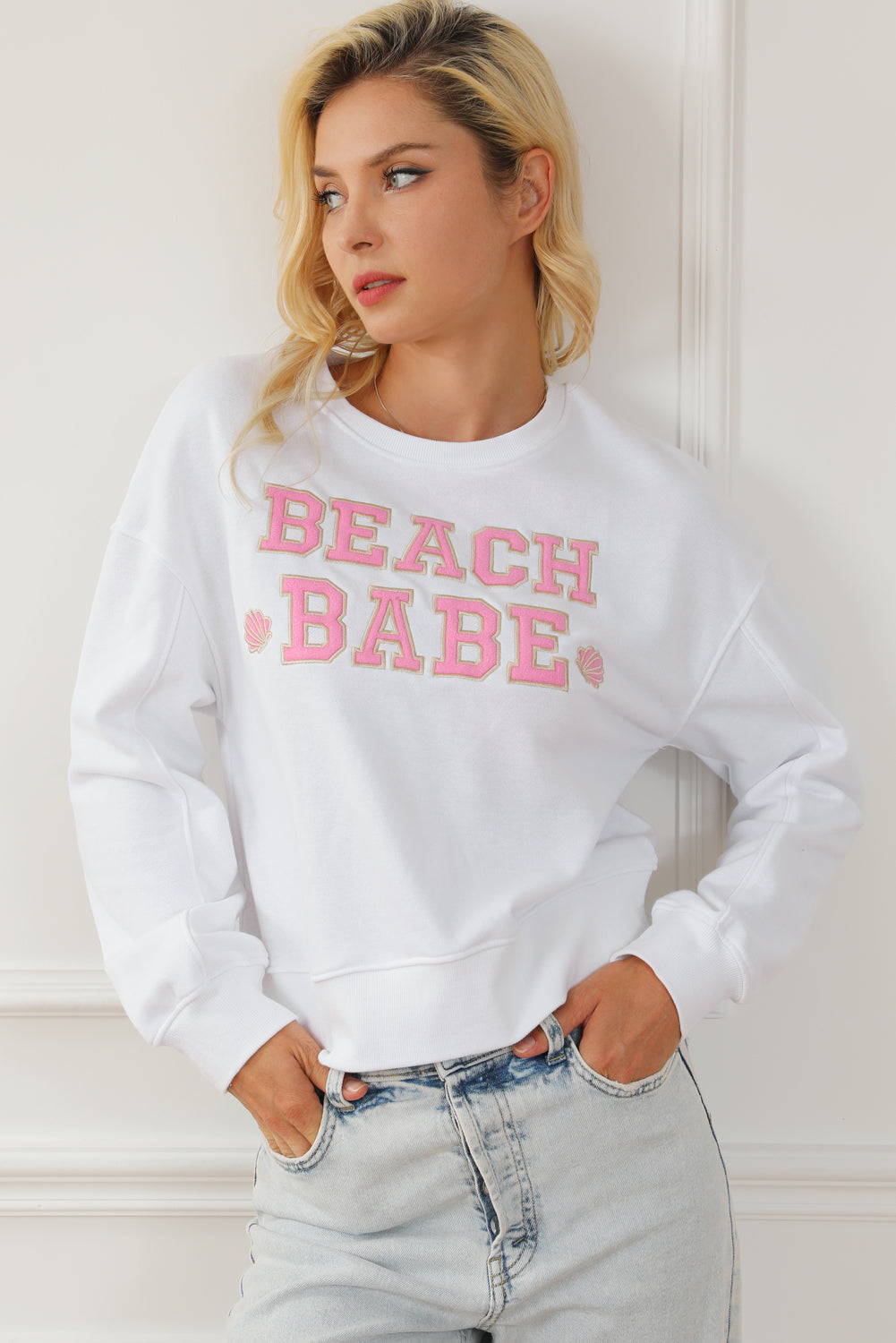 A person with long blonde hair is standing with their back to the camera, wearing a White BEACH BABE Slogan Graphic Casual Sweatshirt and light blue jeans.