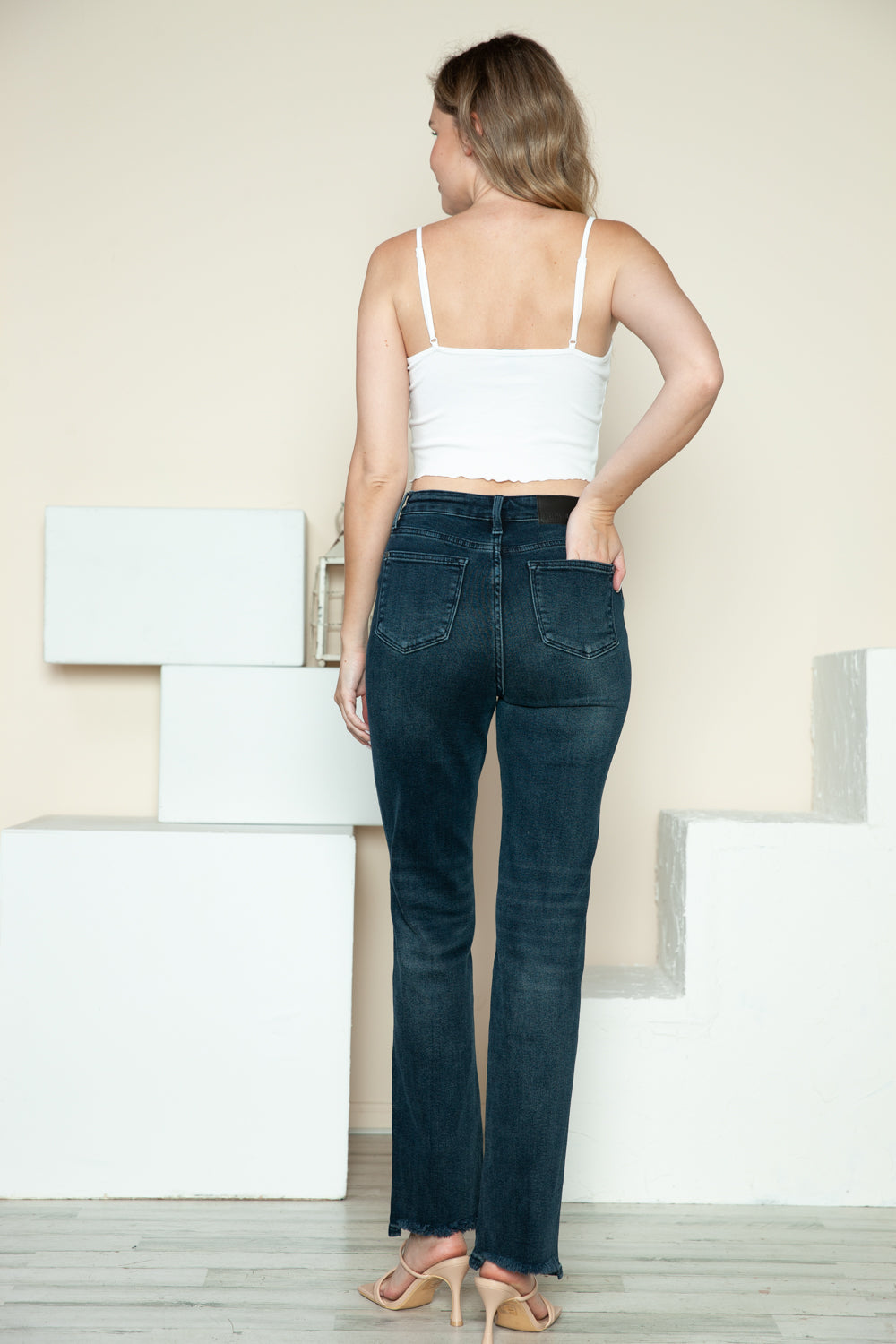 A person is modeling the Judy Blue Full Size Button Fly Hem Destroy Straight Jeans, high-waisted with distressed detailing and frayed hems, paired with light-colored open-toe heels. The background includes white blocks and a light-colored wall.