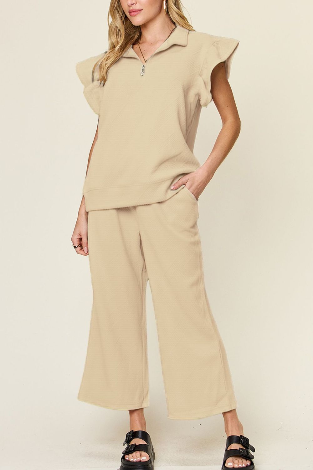 A woman stands wearing the Double Take Texture Ruffle Short Sleeve Top and Drawstring Wide Leg Pants Set in a slightly stretchy, light gray fabric with zipper detail. The two-piece outfit is complemented by black sandals, and she has one hand in her pocket. Machine wash cold for easy care.