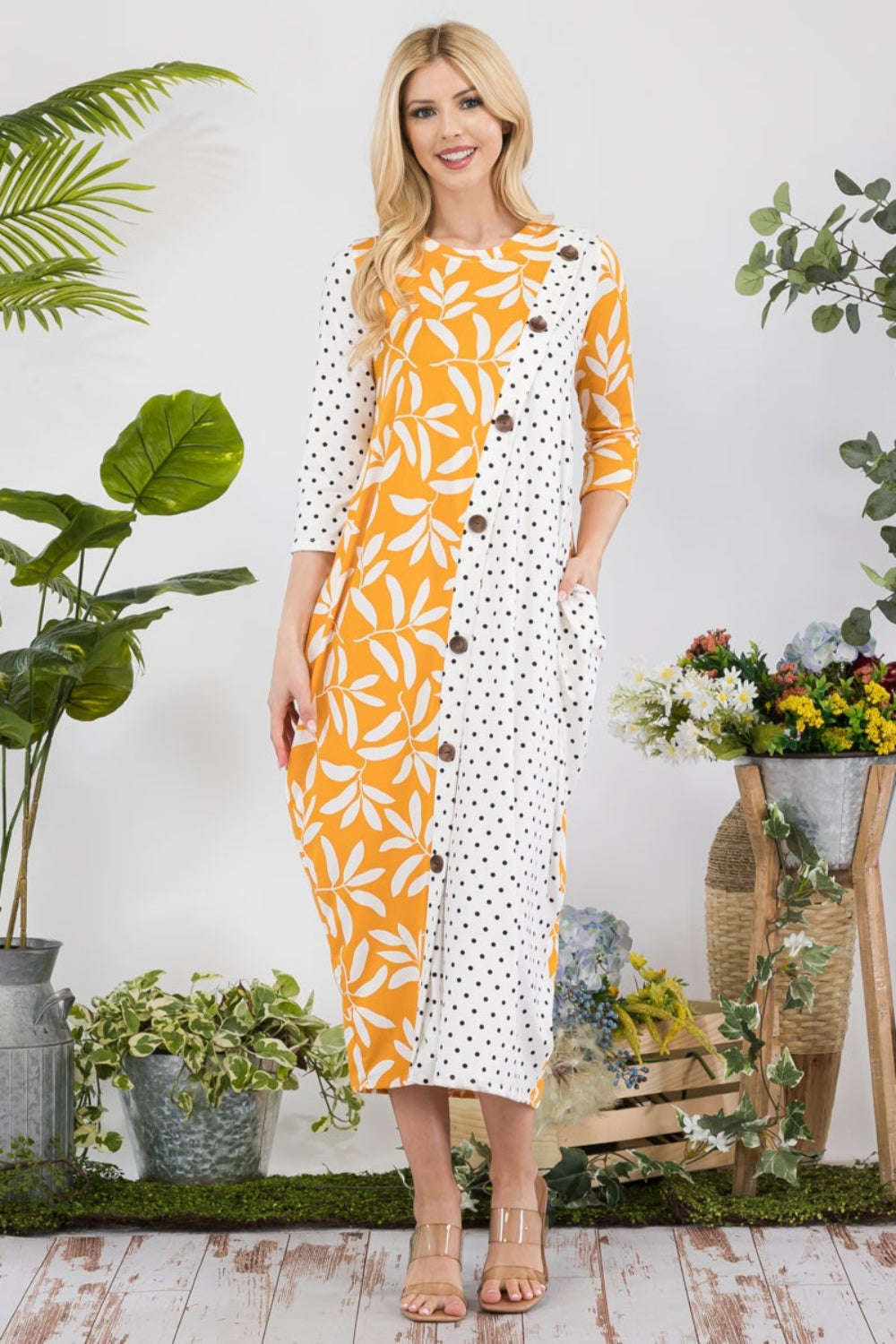 In a garden setting, a woman models the Celeste Full Size Floral Polka Dot Contrast Midi-Dress with Pockets, featuring its versatile yellow design among lush plants and vibrant blooms.