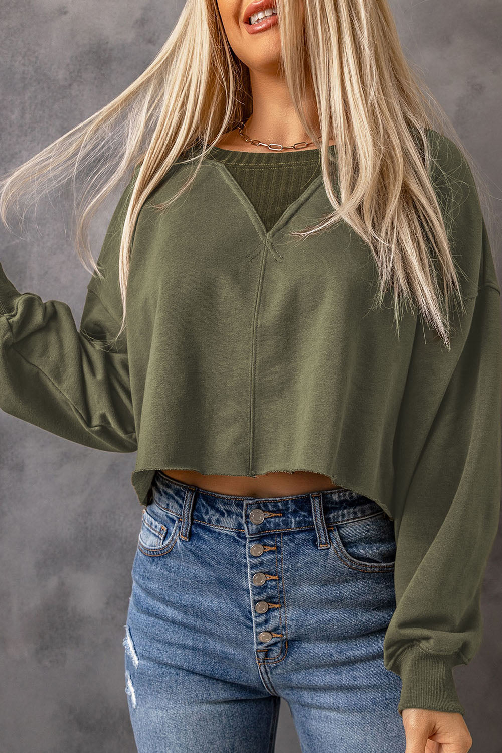 A person with long blonde hair stands with their back to the camera, wearing a faddish and cozy Green Drop Shoulder Cropped Sweatshirt and blue jeans.