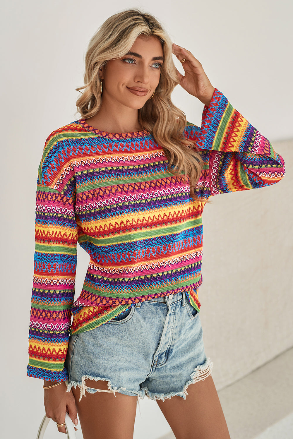 A person stands with hands in the pockets of their denim shorts, wearing a colorful Rose Red Ethnic Wavy Pattern Drop Shoulder Knit Top and a white shoulder bag.