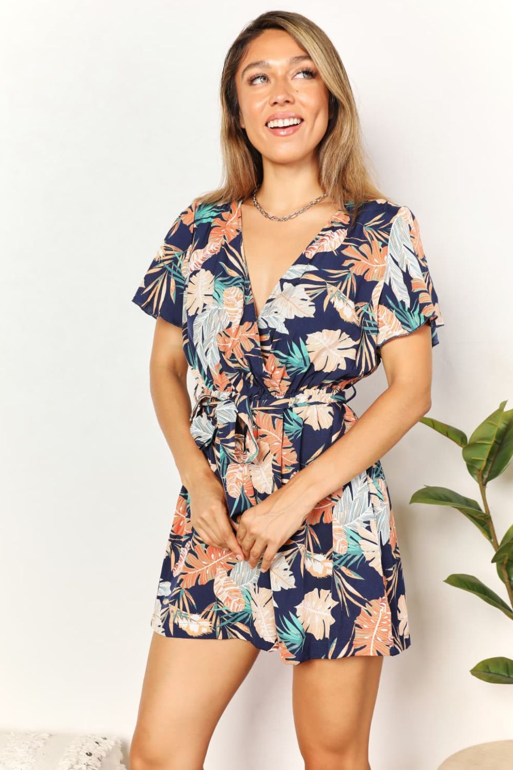 A woman stands smiling with her hands clasped, wearing the Honey Botanical Print Surplice Neck Tie Waist Romper.