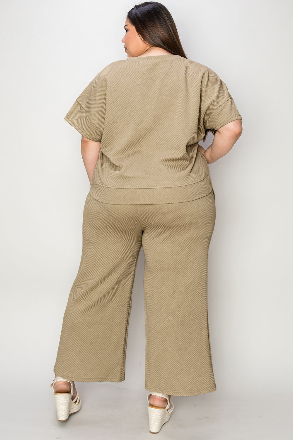 A woman with long hair smiles while standing outside a building, dressed in the Double Take Full Size Texture Short Sleeve Top and Pants Set, featuring a matching beige two-piece ensemble. She wears slides and holds a hat in her right hand. Her outfit, slightly stretchy for added comfort, is perfect for machine wash cold.