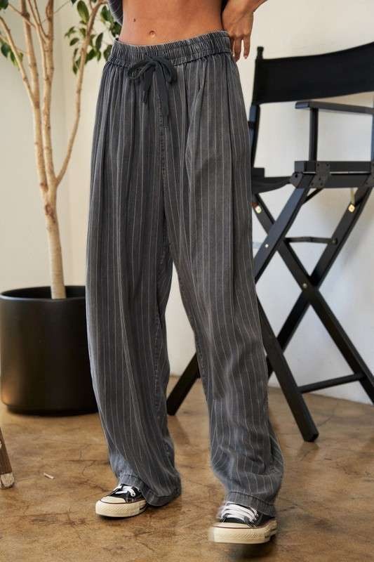 A person wears a casual outfit called the Stripe Button Down Shirt and Long Pants Set, which features a loose-fitting pinstriped design, paired with sneakers. They stand indoors near a potted plant and a director's chair.