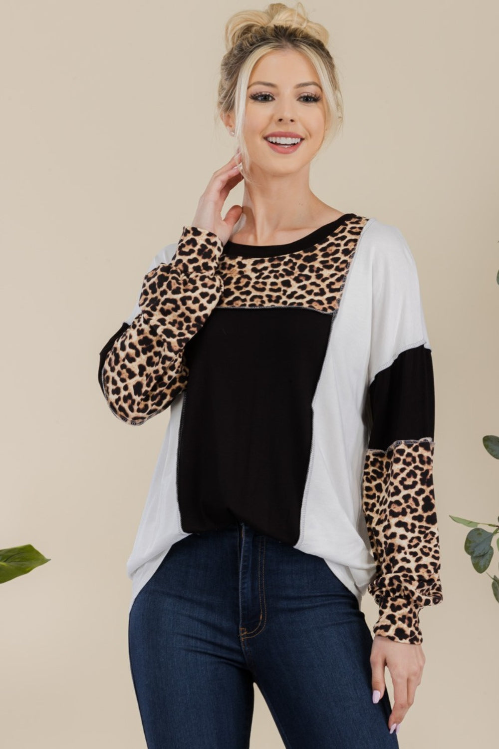 A woman stands in front of a simple background with a plant, wearing the Celeste Full Size Leopard Color Block Exposed Seam T-Shirt, which boasts leopard print cuffs and striking black and white color blocks.