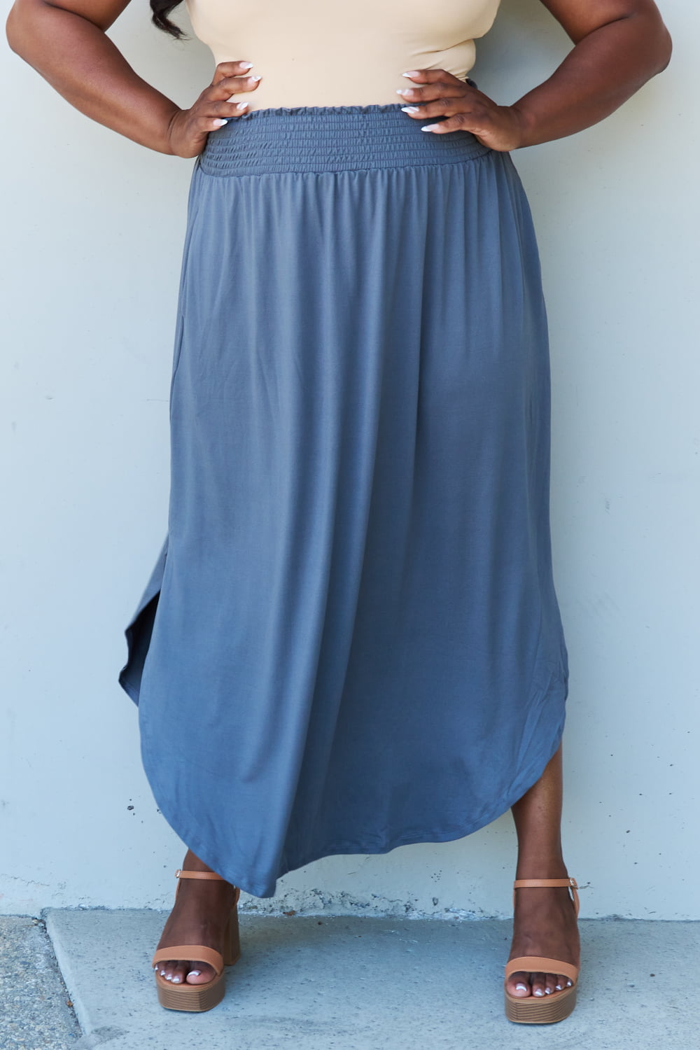 A person wearing a white sleeveless top and the Doublju Comfort Princess Full Size High Waist Scoop Hem Maxi Skirt in Charcoal stands against a plain wall. They are also wearing black sandals.