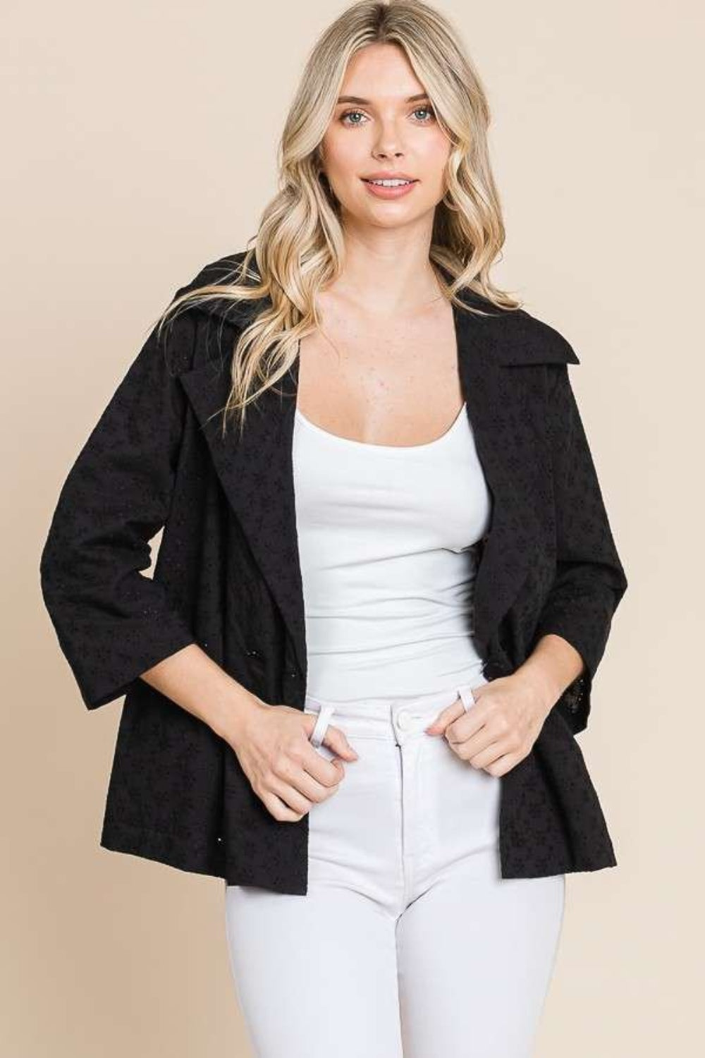 A woman in a chic Culture Code Double Breasted Eyelet Jacket with Pockets and a white top stands gracefully against a beige background.