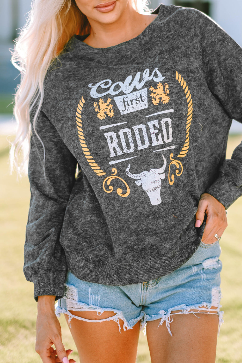 A blonde woman wearing a Gray Coors Banquet RODEO Graphic Mineral Washed Sweatshirt and light blue denim shorts, showcasing casual outdoor wear, stands outdoors with her back to the camera.